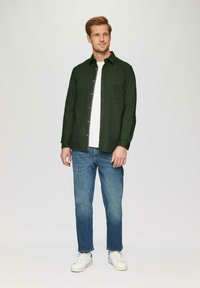 Donkergroene overhemd met knopen en een borstzak, gecombineerd met blauwe jeans en witte sneakers, staande tegen een lichte achtergrond.
