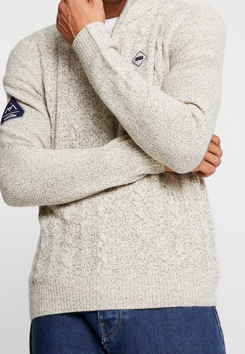 Homme portant un pull en maille torsadée crème avec deux écussons de logo, un sur la poitrine et un sur le haut du bras, assorti à un jean bleu.