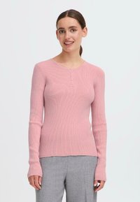 Lyserød ribbet langærmet sweater med rund halsudskæring, der har fem knapper foran og tætsiddende manchetter. Båret over grå bukser.