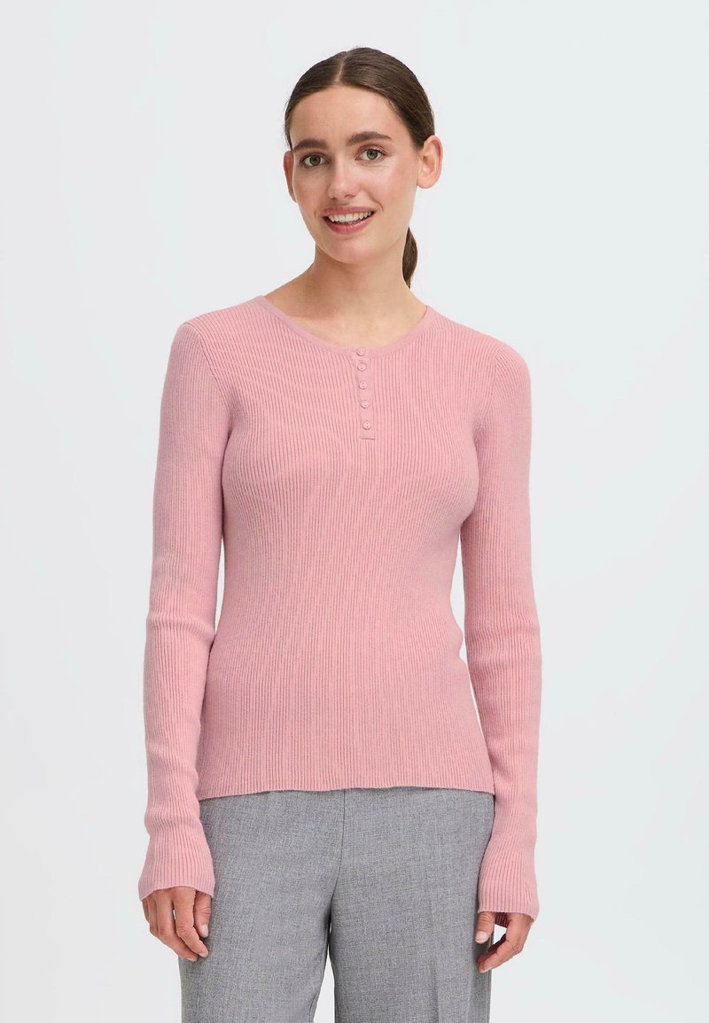 Lyserød ribbet langærmet sweater med rund halsudskæring, der har fem knapper foran og tætsiddende manchetter. Båret over grå bukser.