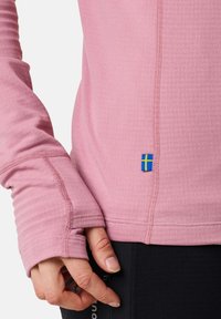 Chemise à manches longues rose clair avec texture côtelée, coutures plates et une petite étiquette avec le drapeau suédois bleu et jaune sur l'ourlet latéral.