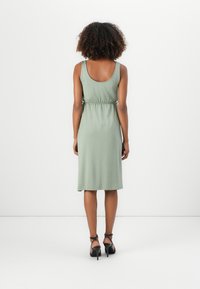 Mulher com cabelo encaracolado vestindo um vestido midi sem mangas de cor verde-sálvia e saltos com tiras pretas, em pé de frente para uma parede branca.