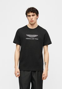 Νεαρός άνδρας με σγουρά μαλλιά που φοράει μαύρο t-shirt της ομάδας Aston Martin Formula One και μαύρο παντελόνι, στέκεται μπροστά σε απλό φόντο.