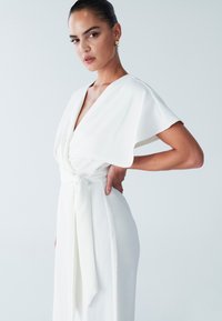 Witte jurk met een diepe V-hals, korte cape-mouwen en een tailledetail met strik. Gladde stof met een vloeiend ontwerp. Subtiele, elegante silhouet.