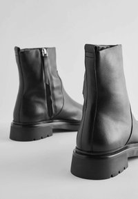 Bottines noires en cuir avec une texture lisse, fermeture éclair latérale et semelles en caoutchouc robustes. Présente une forme incurvée avec un minimum de coutures.