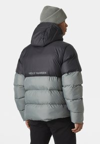 Pufferjack in zwart en lichtgrijs met een capuchon. Beschikt over horizontaal gestikte secties en het "HELly HANSEN" logo op de rug. Geweven gebreide beanie.