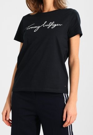 Frau trägt ein schwarzes Tommy Hilfiger T-Shirt mit weißem Logo-Schriftzug und schwarze Hose mit weißen Seitenstreifen, steht vor hellem Hintergrund.