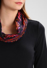 Una sciarpa infinita con motivo rosso e nero presenta vortici e forme geometriche, realizzata in un tessuto morbido, indossata sopra una maglietta scura a maniche lunghe.