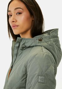 Grüne gesteppte Jacke mit Kapuze, ausgestattet mit einem dunklen Reißverschluss und einem braunen Leder-Ziehband. Enthält ein Logo-Patch auf dem linken Ärmel.