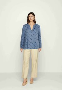Blusa azul com decote em V e mangas longas, apresentando um padrão de contornos escuros, combinada com calças largas de cor creme.