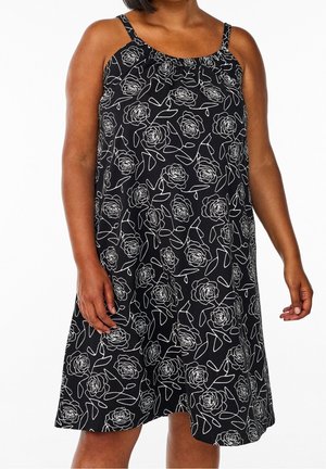 Frau trägt ein schwarzes ärmelloses Kleid mit weißen Umrissen von Rosen und Blättern, steht vor einem einfarbigen weißen Hintergrund.