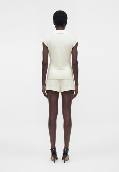 Γυναίκα με κοντά μαλλιά που φοράει λευκό κορυφαίο με κοντά μανίκια και γιακά, ταιριαστά σορτς και μαύρα σανδάλια με ψηλό τακούνι, στέκεται με την πλάτη προς τα έξω.