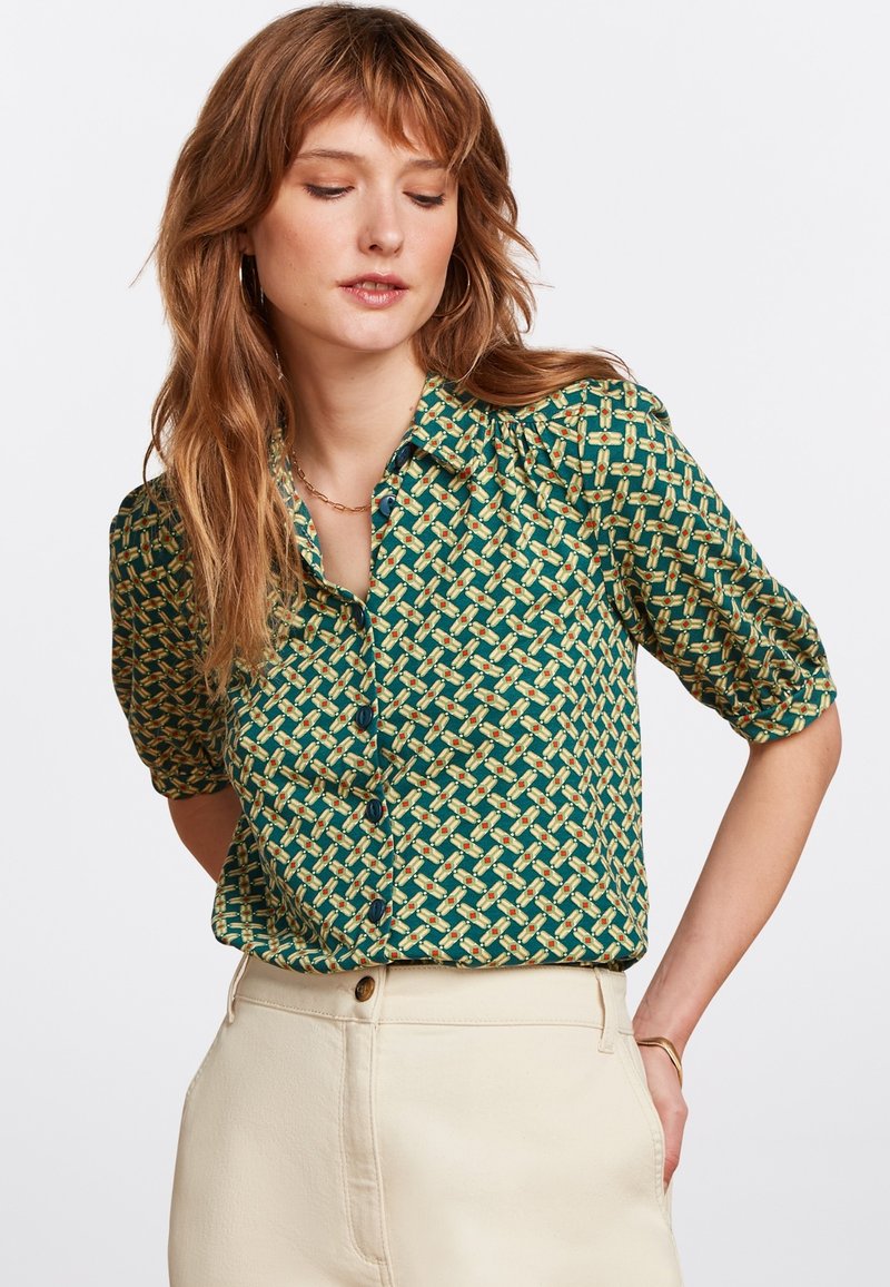 Blusa verde a maniche corte con motivo geometrico giallo e arancione, con chiusura a bottoni frontale e tessuto morbido e leggero.