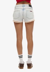 Pantalones cortos de mezclilla azul claro con un dobladillo desgastado y un borde de encaje, con dos bolsillos traseros y una etiqueta negra en la cinturilla. Llevados con calcetines a rayas.