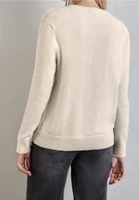 Beige tröja i mjukt stickat tyg, med ribbat avslut och manschetter, klassisk rund halsringning och skräddarsydd passform, kombinerad med mörka jeans.