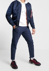 Granatowa kurtka bombowa z ribbowanymi mankietami, pod spodem biała bluza z kapturem, dopasowane ciemnoniebieskie spodnie oraz czarne sneakersy z burgundowymi akcentami.