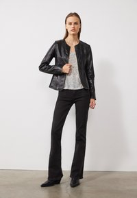 Giacca in ecopelle nera con collo rotondo, chiusura con zip e superficie liscia, abbinata a un top grigio e jeans neri a campana.