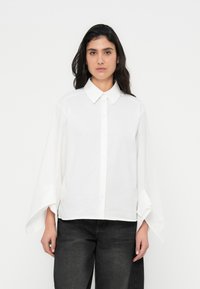 Λευκό πουκάμισο με κουμπιά, γιακά και φαρδιές, φουσκωτές μανσέτες. Κατασκευασμένο από απαλό ύφασμα με χαλαρή εφαρμογή. Συνδυασμένο με σκούρο τζιν.