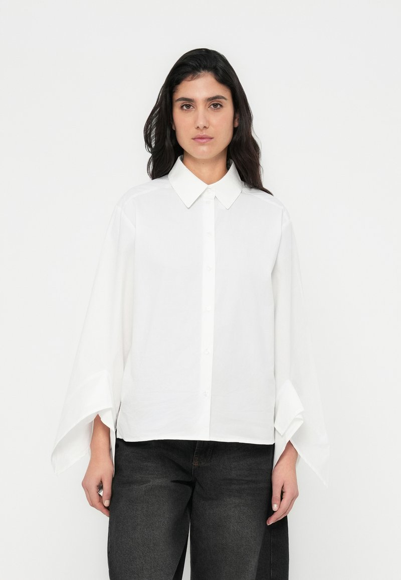 Λευκό πουκάμισο με κουμπιά, γιακά και φαρδιές, φουσκωτές μανσέτες. Κατασκευασμένο από απαλό ύφασμα με χαλαρή εφαρμογή. Συνδυασμένο με σκούρο τζιν.