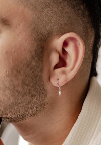 Orecchino a cerchio in argento con una piccola goccia di perla, attaccato a un orecchio. L'angolo ravvicinato rivela una texture liscia e una finitura lucida.