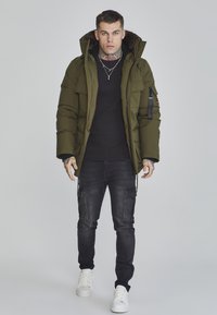 Puffetjakke i olivengrøn med hætte, lynlås foran, to forlommer og en sort ribstrikket sweater under. Mørke jeans og hvide sneakers.