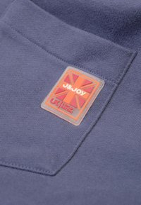 Donkerblauwe sweatshirtstof met een gestikte zak. Bevat een rubberen label met de afbeelding van een Union Jack en de tekst "J&JOY" in oranje en wit.
