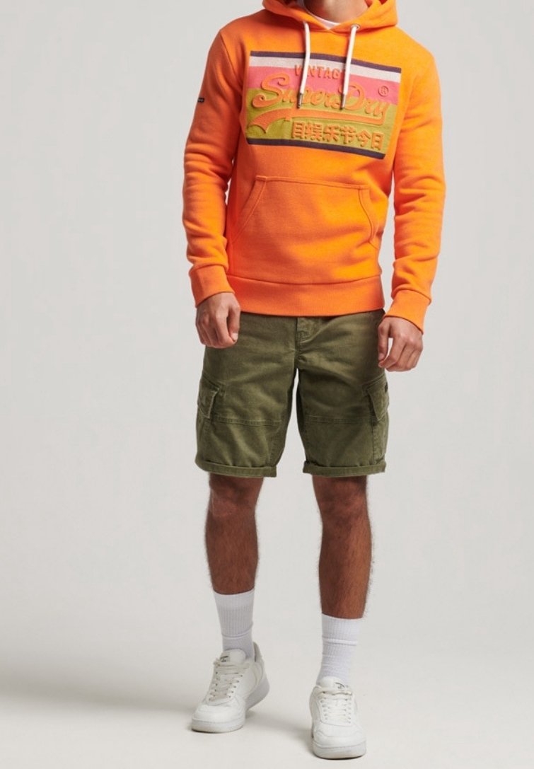 Homme portant un sweat à capuche orange vif avec un logo coloré, un short cargo vert olive, des chaussettes blanches et des baskets blanches, debout devant un fond uni.