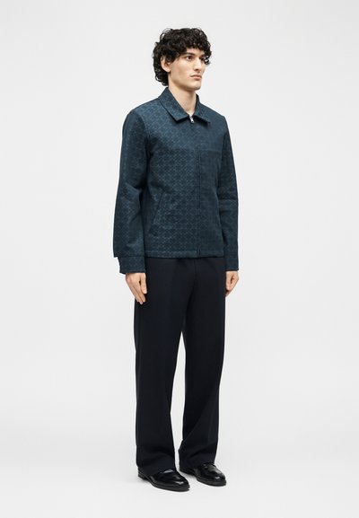 Άνδρας με σγουρά μαλλιά που φοράει σκούρο μπουφάν με φερμουάρ και σχέδια, μαύρο παντελόνι και μαύρα δερμάτινα παπούτσια, στέκεται μπροστά σε λευκό φόντο.