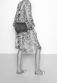 Czarno-białe zdjęcie przedstawiające osobę w wzorzystej, luźnej sukience i czarnej torbie na ramię z srebrną klamrą, do tego płaskie sandały.