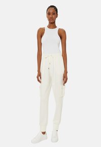 Top bianco senza maniche abbinato a pantaloni jogger crema con tasche cargo, vita con cordino e polsini a coste. Il modello indossa sneakers bianche.