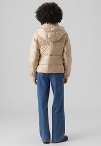 Beige puffer-takki hupulla, jossa tikattu kuvio ja näkyvät saumat. Yhdistetty sinisiin leveälahkeisiin farkkuihin ja mustiin kenkiin.