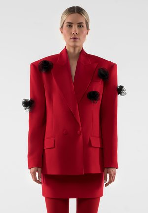 Kobieta w oversize'owym czerwonym blezerze z usztywnionymi ramionami, ozdobionym czarnymi aplikacjami kwiatowymi, stojąca na jednolitym tle.