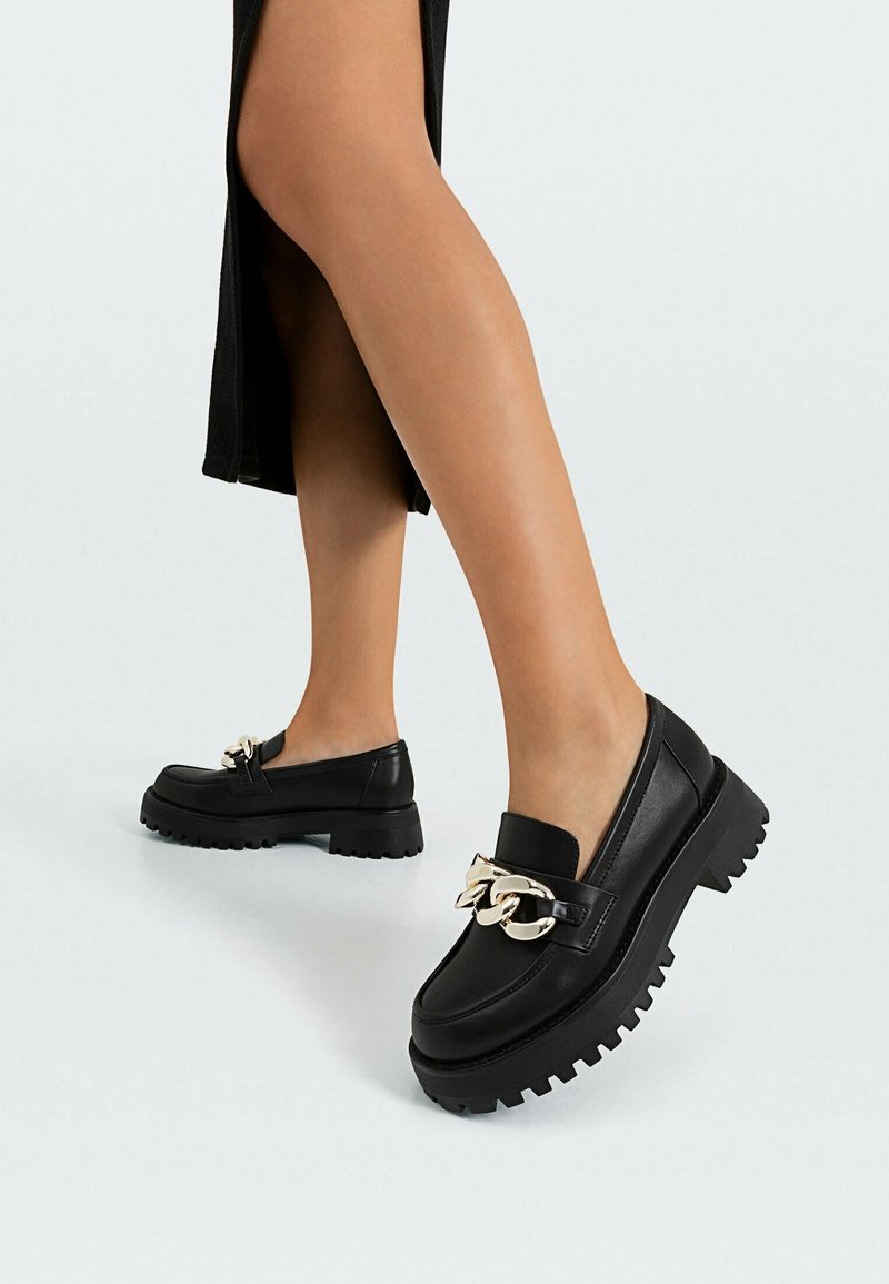 Mocassins en cuir noir avec une semelle épaisse, ornés d'une chaîne dorée proéminente. Texture lisse et design à bout arrondi.