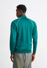 Jachetă sport verde cu guler înalt, cu detalii galbene pe umeri și mâneci, realizată dintr-un material neted și elastic.