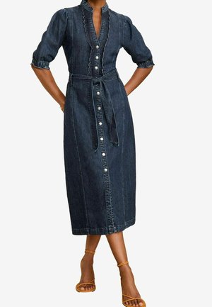 Vrouw draagt een donkerblauwe midi denimjurk met pofmouwen, knoopsluiting aan de voorkant, gegolfde randen en een tailleriem met strik, gecombineerd met tankleurige sandaalhakken met bandjes.