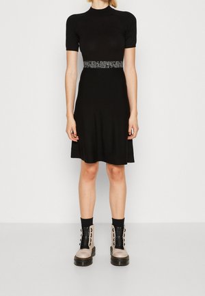 Femme portant une robe noire à manches courtes avec une ceinture à motif à la taille et des bottines beige clair avec des lacets et des semelles noires.