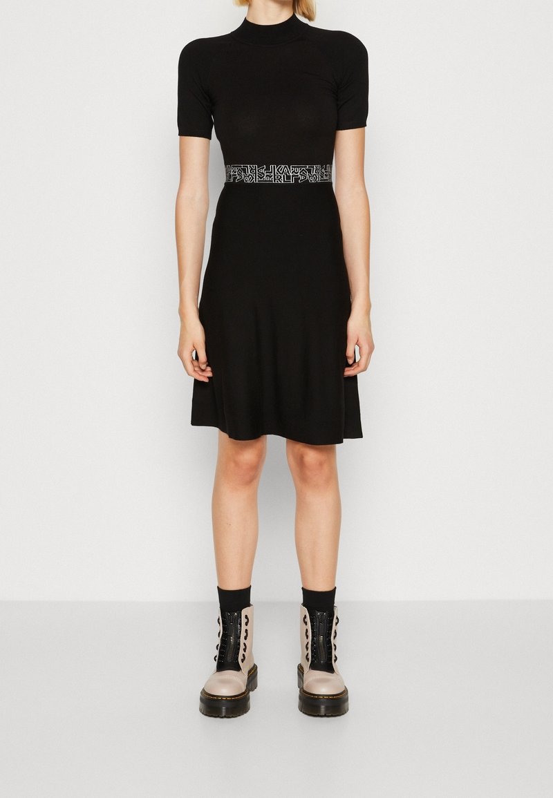 Femme portant une robe noire à manches courtes avec une ceinture à motif à la taille et des bottines beige clair avec des lacets et des semelles noires.