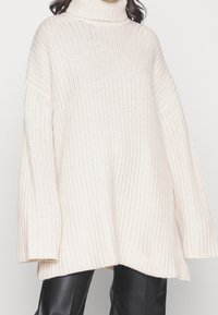 Vrouw in een oversized, crèmekleurige ribgebreide coltrui, gecombineerd met zwarte leren broek, tegen een effen achtergrond.