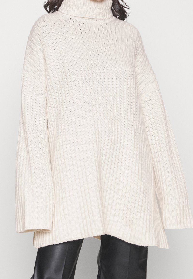 Vrouw in een oversized, crèmekleurige ribgebreide coltrui, gecombineerd met zwarte leren broek, tegen een effen achtergrond.