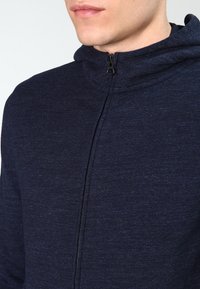 Mörkblå zip-up huvtröja med hög krage, med en slät yta och subtilt melerat mönster. Dragkedjestängning fram.