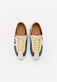 Lyse grå sneakers med marineblå detaljer, gule snøringer og brune såler. Har kontrasterende sømmer og et strømlinjeformet design.