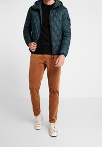 Turkos vadderad jacka med huva över en svart texturerad tröja, kombinerad med bruna kordfloretbyxor och beige sneakers. En enkel och avslappnad outfit.