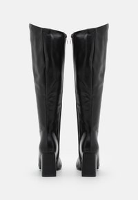 Paire de bottes noires en cuir hautes avec des talons carrés et des fermetures éclair à l'arrière, présentées de l'arrière sur un fond blanc.