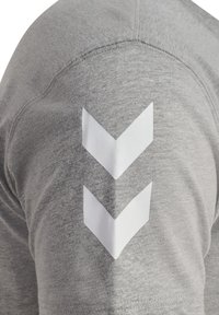 Grå kortärmad skjorta med vita chevrongrafik på vänster axel; mjukt material med synliga sömmdetaljer.