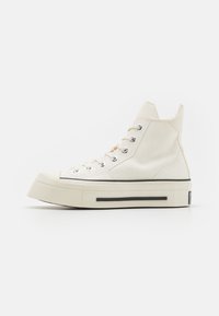 Adidași tip high-top din pânză de culoare off-white, cu ochiuri argintii, talpă groasă din cauciuc crem și o dungi accent neagră de-a lungul bazei.