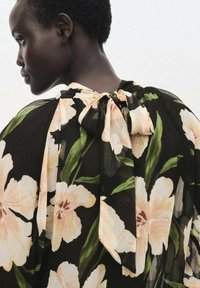 Blusa nera a fiori con grandi fiori rosa chiaro e foglie verdi. Presenta un nastro da legare sul retro e un tessuto fluido e trasparente.
