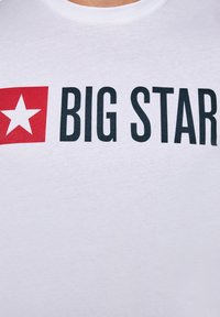 Wit shirt met een rood vierkant waarin een witte ster staat, naast vetgedrukte zwarte tekst "BIG STAR" over de borst.