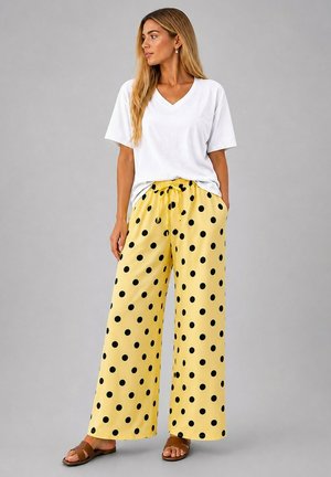 Femme portant un pantalon jaune ample à pois noirs, un t-shirt blanc à manches courtes col en V et des sandales marron, debout devant un fond gris uni.