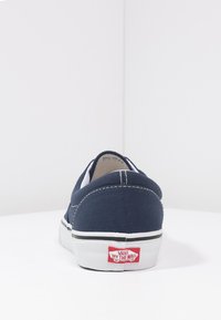Mørkeblå kanvas slip-on sko med hvit gummisåle og svart stripe. Har hvite sømmer og rød "Vans Off The Wall"-logo på hælen.