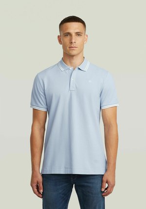 Young man with short hair wearing a light blue polo shirt with white trim and dark blue jeans, standing against a neutral background.